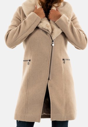 Kobieta poprawiająca beżowy wełniany płaszcz z dużym futrzanym kołnierzem, przednim zamkiem błyskawicznym i zasuwanymi kieszeniami po bokach, pod spodem nosi ciemne spodnie.