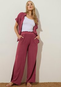 Losse, roze stoffen broek met zijsplits, gecombineerd met een wit shirt en een bijpassend jasje. Sandalen maken de outfit compleet.