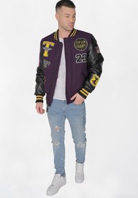 Violet yliopistojakki mustilla nahkahihnoilla, keltaisia yksityiskohtia ja erilaisia applikaatioita. Yhdistetty käytettyihin vaaleansinisiin farkkuihin ja valkoisiin kenkiin.