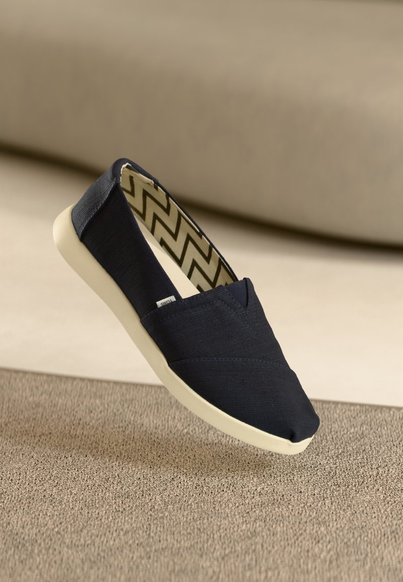 Chaussure en toile bleu marine sans lacets avec semelle blanche et doublure intérieure à motif zigzag noir et blanc reposant sur une surface textile beige.