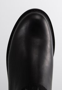 Černá kožená kotníková bota s hladkým povrchem, zaoblenou špičkou a jemným prošíváním podél okraje. Obsahuje texturovanou tkaninovou část na straně.