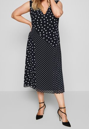 Mujer con un vestido midi sin mangas negro con diferentes patrones de lunares blancos, combinado con tacones altos negros con tiras.
