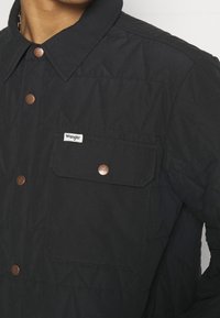 Black quilted jacket with a fold-over collar, copper snap buttons, and a chest pocket featuring a small white brand label.