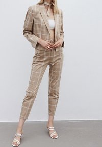 Traje de cuadros beige con un blazer a medida y pantalones ajustados. El tejido parece suave; combinado con calzado blanco y un detalle en el cuello a juego.