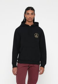 Hoodie preto com um bolso frontal e cordão, apresentando um logótipo dourado no peito. O modelo usa calças maroon.