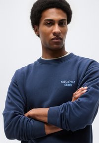 Άντρας με σταυρωμένα χέρια, φορώντας μπλέναζι φούτερ Marc O'Polo Denim, κοιτάζοντας με αυτοπεποίθηση μπροστά, σε απλό ανοιχτόχρωμο φόντο.