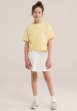 Jong meisje staand, gekleed in een geel shirt met korte mouwen met kleine bloemmotieven, een witte rok, witte sokken en zilveren sneakers.