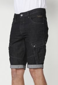 Schwarze Denim-Cargoshorts mit umgeschlagenem Saum, ausgestattet mit Seitentaschen und Stitching-Details. Klassische Passform mit Knopfverschluss und Gürtelschlaufen.