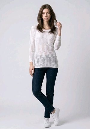 Mujer de pie sobre un fondo liso, vistiendo un jersey blanco de punto de manga larga, vaqueros oscuros y zapatillas blancas, con una mano tocándose el cabello.