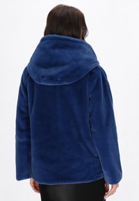 Giacca con cappuccio in peluche blu navy dal morbido texture, forma squadrata e spalle scese. Nessun hardware o motivo visibile.