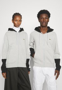 Felpa grigia con zip e accenti neri, dotata di cappuccio. Presenta strisce laterali bianche a contraste e un piccolo logo verde sul petto.