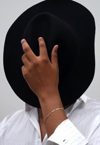 Black felt hat held by a hand with a gold chain bracelet, paired with a white striped shirt. Focus on the contrast of materials and textures.