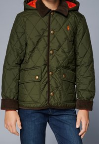 Vihreä tikattu takki, jossa on ruskea corduroy-kaulus, oranssi vuori ja etutaskut. Takissa on kultaiset napit ja kiristysnauhakapussi.