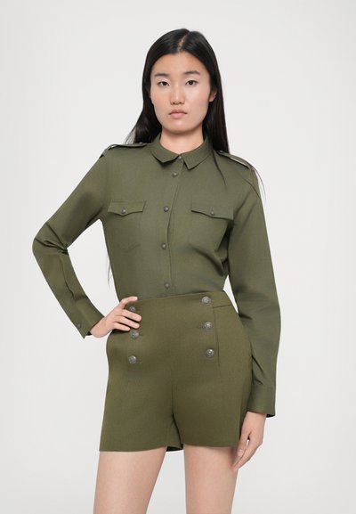 Olivovo zelená vojenská košeľa s dlhými rukávmi, zapínaním na gombíky vpredu a dvoma vreckami na hrudi, kombinovaná s kraťasmi s vysokým pásom, ktoré majú dvojitý gombíkový zapínanie.