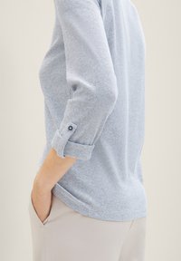Camisa de rayas azul claro con mangas tres cuartos, puños enrollados y un tejido texturizado, combinada con pantalones de color claro.