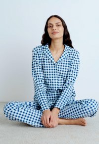 Conjunto de pijama em padrão de xadrez azul e branco, com mangas longas e calças, modelo sentado, mãos juntas, fundo em carpete bege.