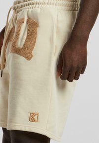 Beige shorts with an elastic waistband, featuring a plush terry cloth "H" detail and a small tan logo patch at the hem. Soft cotton material.