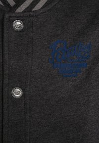 Chaqueta de tela de jersey gris oscuro con cuello a rayas. Presenta un logotipo bordado en azul y detalles en botones metálicos. Material suave y texturizado.