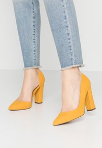 Chaussures à talons hauts jaunes texturées avec un bout pointu et un talon épais, associées à un jean bleu clair avec un ourlet effiloché.