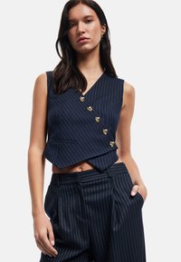 Marineblaues Nadelstreifen-Weste mit V-Ausschnitt, goldenen Knopf-Akzenten und einem cropped Design, kombiniert mit hoch taillierten Nadelstreifen-Hosen.