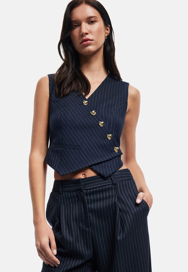 Marineblaues Nadelstreifen-Weste mit V-Ausschnitt, goldenen Knopf-Akzenten und einem cropped Design, kombiniert mit hoch taillierten Nadelstreifen-Hosen.
