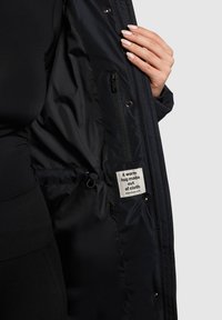 Musta jope sisevooder läikiva nailonmaterjaliga, lukuga tasku, nöörsanga detail ja silt, mis ütleb: „Soe kallistus riidest valmistatud.”