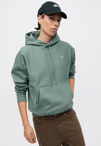Zelená bavlnená mikina s kapucňou a šnúrkou, klokanková vrecko a biele logo Nike, skombinovaná s hnedými nohavicami a čiapkou s logom v čiernej farbe.