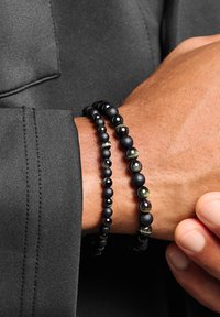 Schwarze Perlenarmbänder; eines mit mattschwarzen Perlen, das andere mit glänzenden schwarzen und grün akzentuierten Perlen, getragen an einem Handgelenk vor dunklem Hintergrund.