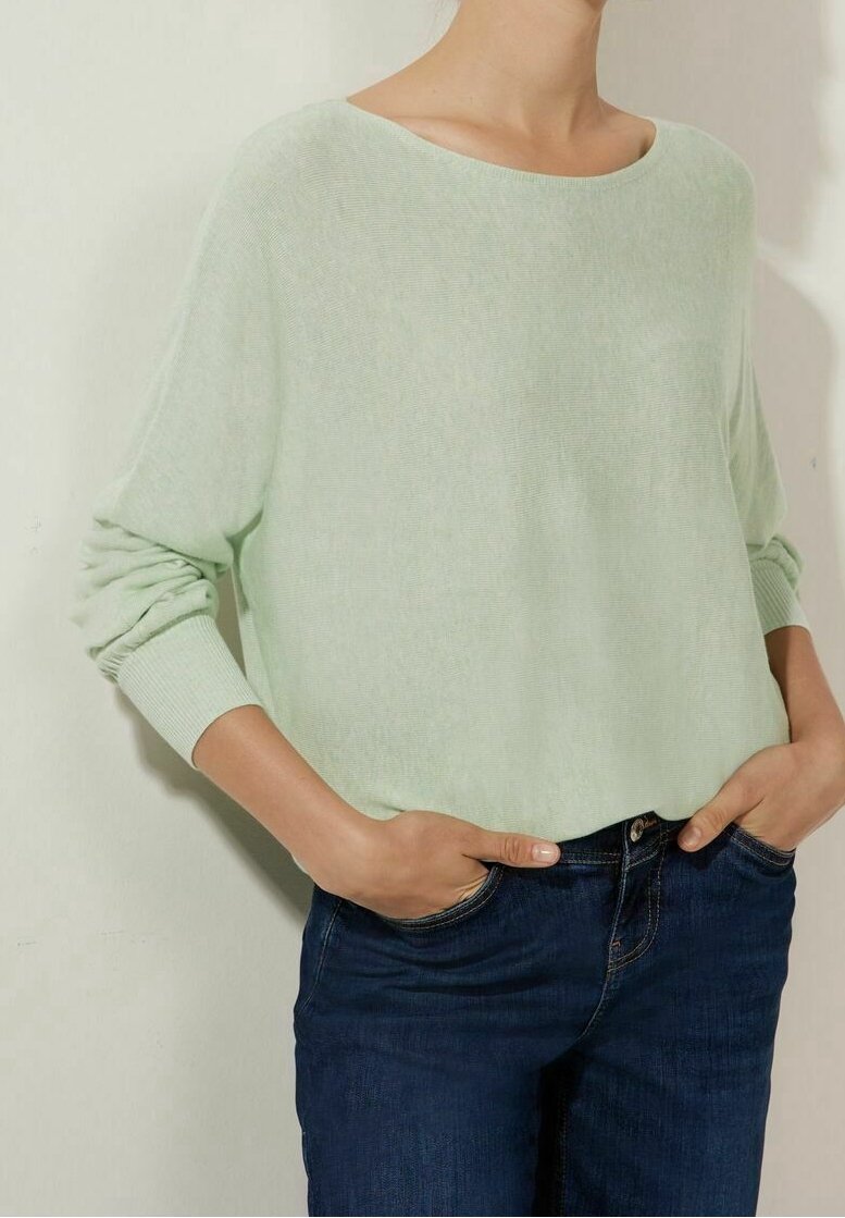 Personne portant un pull vert clair à manches longues et un jean bleu foncé, les mains dans les poches, devant un mur clair uni.
