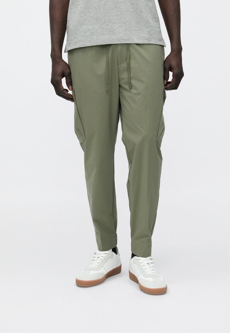 Mees, kes kannab oliivrohelisi pükse paeladega ja valgeid tossusid pruunide talladega, seisab ühevärvilise tausta ees.