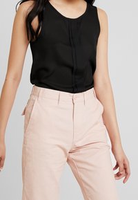 Černý bezrukávový top s vertikálním skladem, kombinovaný se světle růžovými kalhotami s vysokým pasem, které mají boční kapsy a zapínání na knoflík.