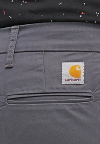 Szare spodnie z bawełny, z tylną kieszenią, żółtą metką z logo Carhartt oraz subtelną teksturą. Widoczny wyszywany wykończenie na brzegach.