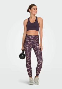 Fioletowy stanik sportowy i legginsy w ciemnych wzorach z złotymi akcentami; w słuchawkach, trzyma czarny kettlebell.