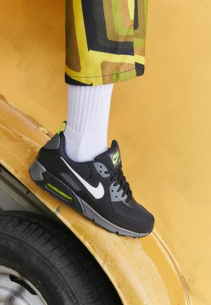 En person som bär svarta och grå Nike Air Max-sneakers med vita strumpor och mönstrade byxor står på en gul bils skärm nära ett däck.