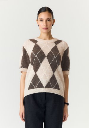 Frau mit dunklen Haaren, die einen beige-braun karierten kurzärmeligen Pullover und schwarze Hose trägt, steht vor einem einfarbigen Hintergrund.