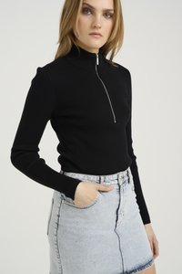 Maglione nero a coste con chiusura a mezza zip, abbinato a una minigonna in denim azzurro chiaro. L'outfit presenta textures aderenti e stili casual.