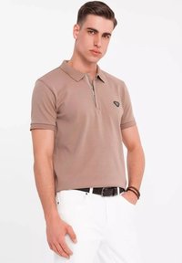 Camisa polo beige con cuello con cremallera, mangas cortas y un acento de logo verde, combinada con pantalones blancos y un cinturón negro. Textura suave.