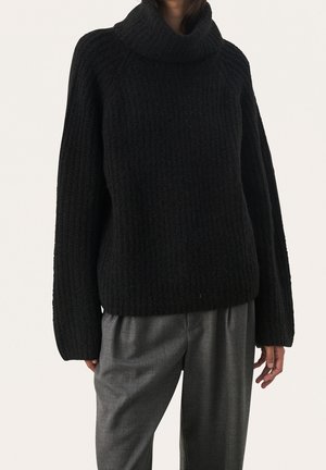 Person wearing a black chunky knit turtleneck sweater and loose gray trousers, standing against a plain light background.