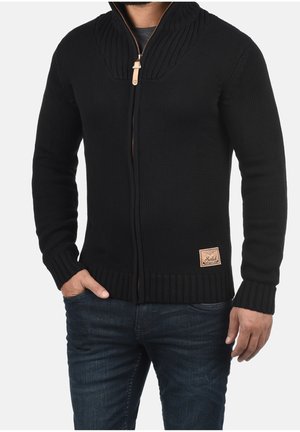 Mand iført en sort strikket lynlåstrøje med ribstrikket krave, manchetter og kant, kombineret med mørkeblå jeans, hånd i lommen.