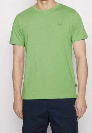 Mężczyzna noszący jasną zieloną koszulkę z krótkim rękawem z małym napisem "JOOP! JEANS" na lewej piersi oraz granatowe spodnie.