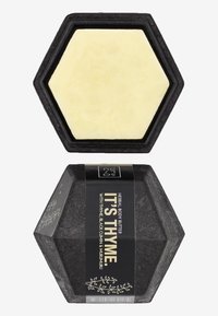 Hexagonal body butter in a black container, with a light yellow product inside. Label features herbal ingredients: thyme, black cumin, murumuru.