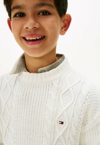 Hvit kabelstrikket genser med ribbestrikket krage og en liten logo aksent. Den indre kragen er i en kontrasterende lysegrå farge.