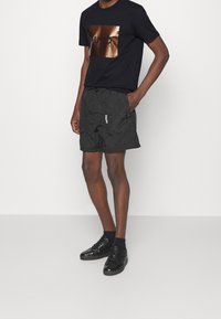 Schwarzes T-Shirt mit glänzendem braunem Logo-Druck, kombiniert mit schwarzen Shorts mit elastischem Bund und weißem Logo. Schwarze Sneaker runden den Look ab.