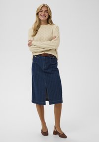 Pull en laine crème avec motif œillet, associé à une jupe en denim bleu marine mi-longue avec des rayures verticales, une fente avant et des poches. Ceinture marron.
