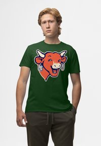 Zelené bavlněné tričko s potiskem usmívající se oranžové krávy s bílými rohy a náušnicemi, zobrazené ve frontálním pohledu.