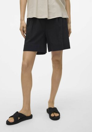 Zwarte katoenen shorts met een relaxte pasvorm en omgeslagen zoom, gecombineerd met zwarte gevlochten slide-sandalen. Achtergrond is eenvoudig wit.