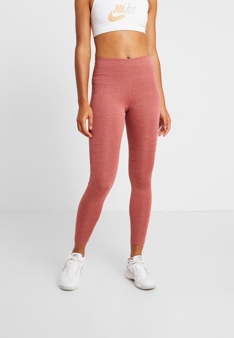 Femme portant des leggings mauve taille haute, un soutien-gorge de sport blanc Nike et des baskets blanches Nike, debout sur un fond blanc uni.