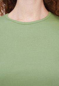 Gros plan d'une personne portant un t-shirt côtelé vert clair à col rond, montrant le cou et la partie supérieure de la poitrine avec des cheveux bouclés aux bords.