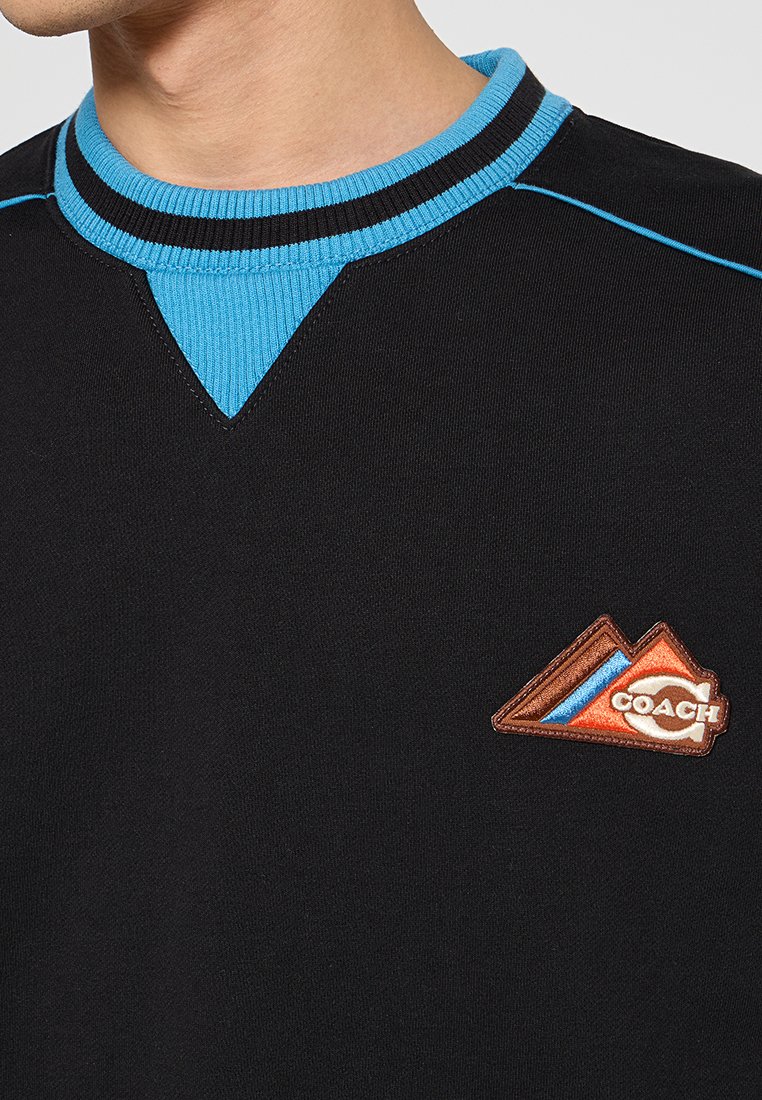 Czarny sweter z dzianiny z niebieską ściągaczem oraz trójkątną brązowo-pomarańczową naszywką z logo "COACH" umieszczoną w widocznym miejscu.