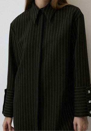 Frau trägt ein schwarzes Nadelstreifen-Langarmhemd mit verlängerten Manschetten und silbernen Knopfdetails, steht vor einem einfarbigen Hintergrund.